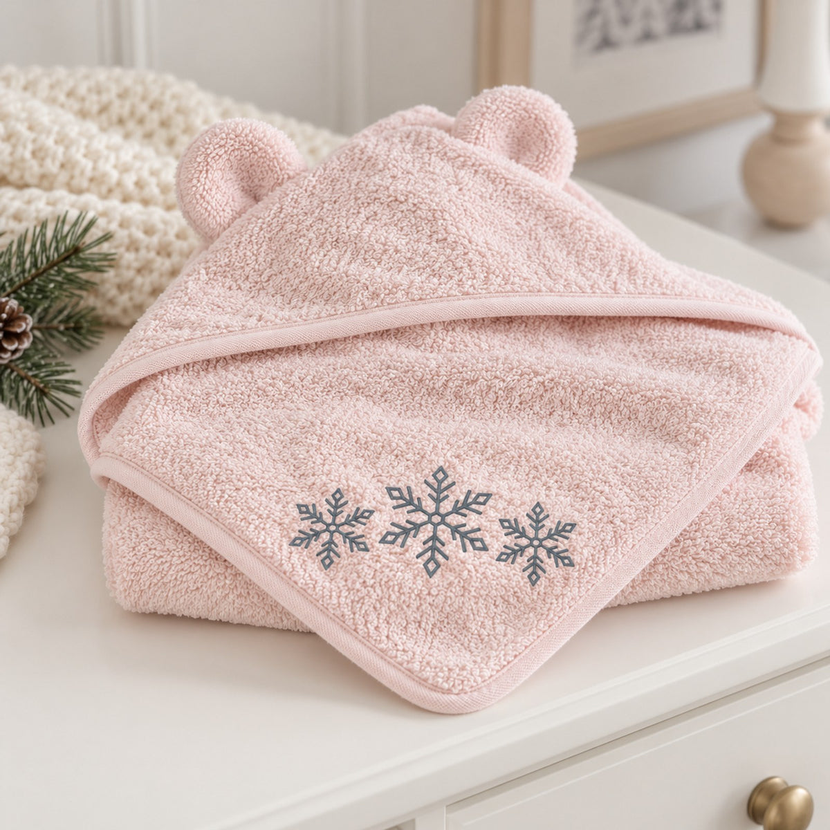 Pink bear-ear baby hooded towel with three embroidered grey snowflakes, folded on a white nursery dresser