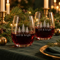 Two stemless wine glasses etched with evergreen trees and still & bright, with red wine on a Christmas table