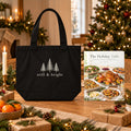The Holiday Table Cookbook & Tote Set: The Holiday Table paperback cookbook by Rachelle Merchant beside the black Still & Bright Evergreen Organic Cotton Tote on a Christmas table