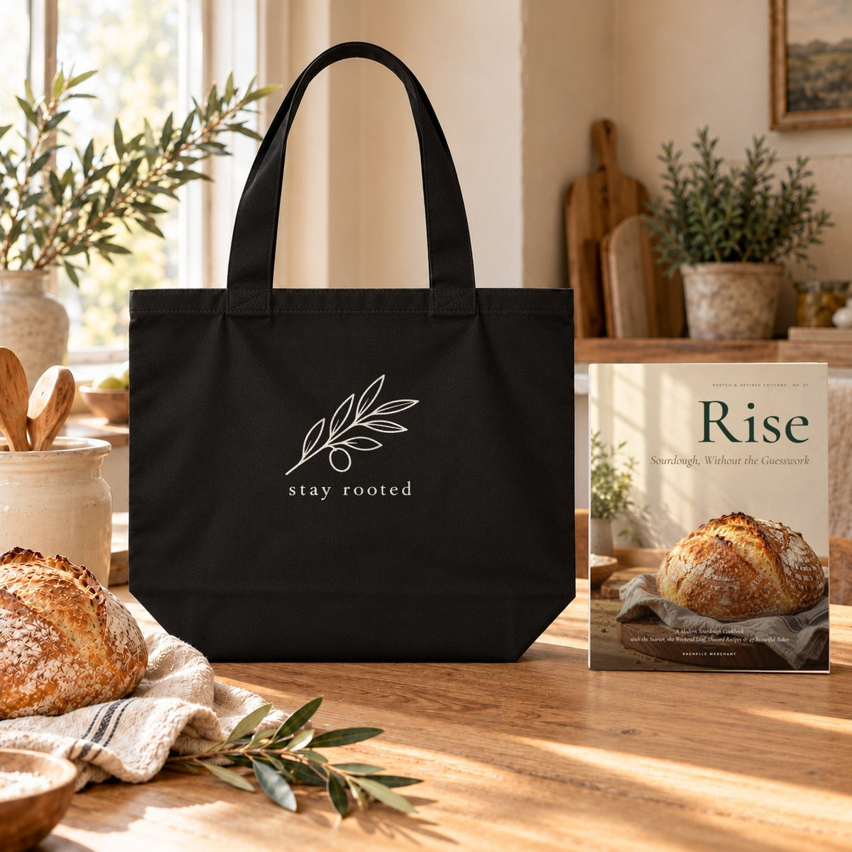 Rise Cookbook & Tote Set: the Rise paperback cookbook by Rachelle Merchant beside the black Stay Rooted Olive Branch Organic Cotton Tote on a kitchen table
