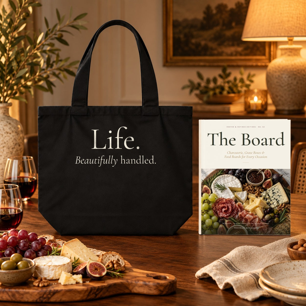The Board Cookbook & Tote Set: The Board paperback cookbook by Rachelle Merchant beside the black Life. Beautifully Handled. Shoulder Tote with a charcuterie spread