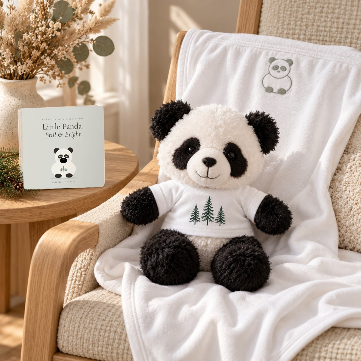 Little Panda Book, Plush & Blanket Gift Set by Rooted & Refined: the Little Panda plush on the white embroidered jersey blanket draped over a nursery rocking chair, with the Little Panda, Still & Bright board book