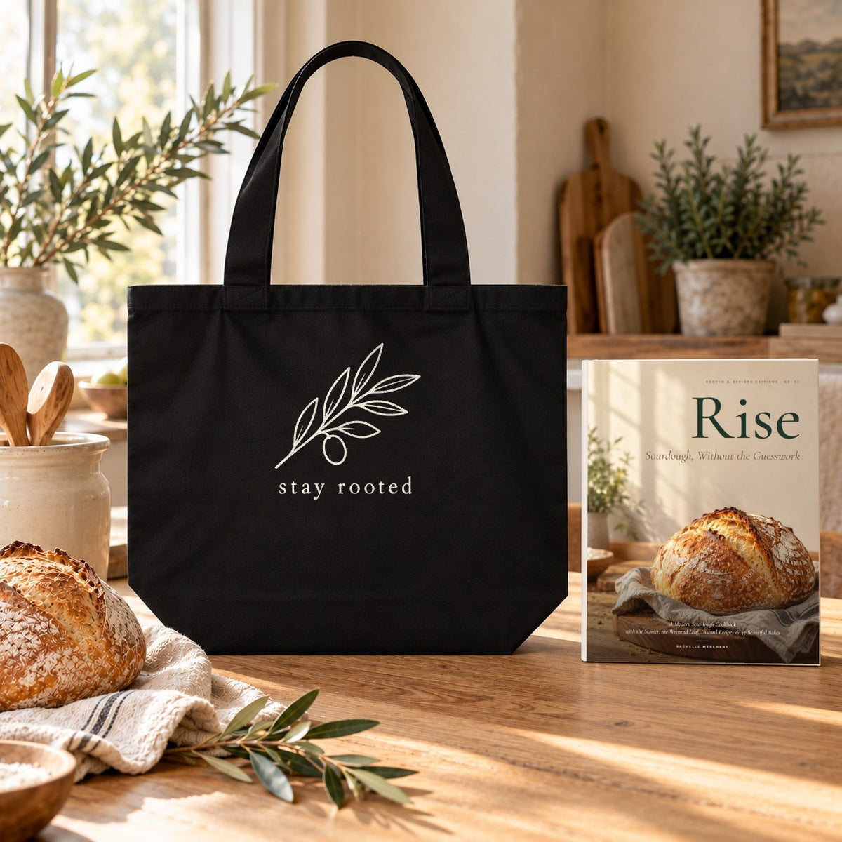 Rise Cookbook & Tote Set: the Rise paperback cookbook by Rachelle Merchant beside the black Stay Rooted Olive Branch Shoulder Tote on a kitchen table