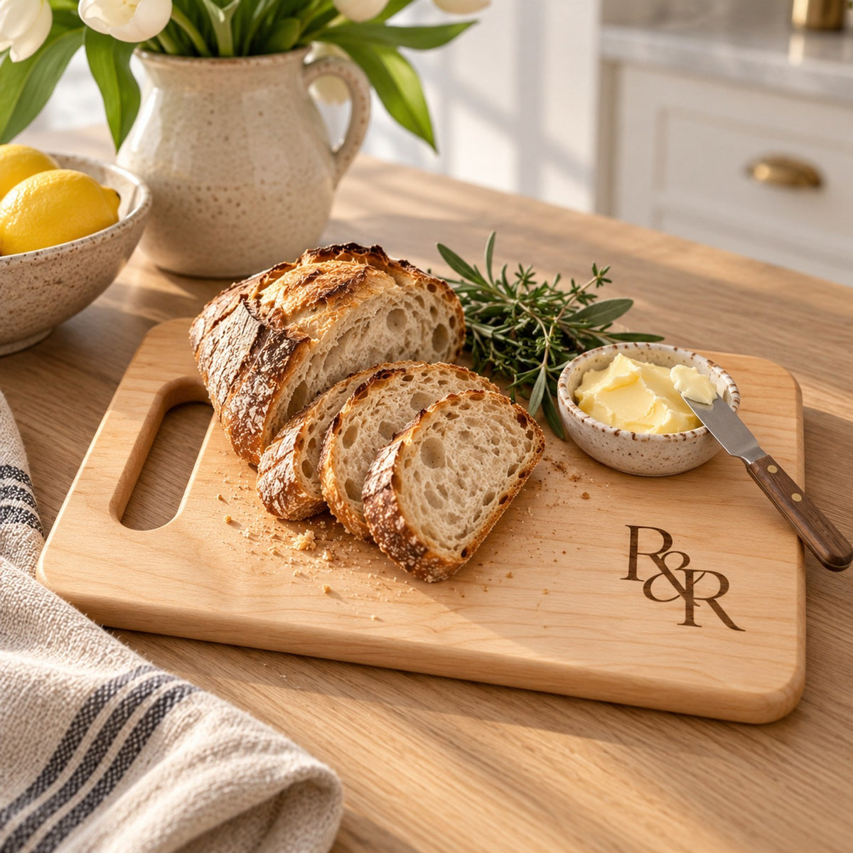Maple cutting board engraved with the R&R monogram, with sliced sourdough, butter and rosemary on a bright kitchen counter
