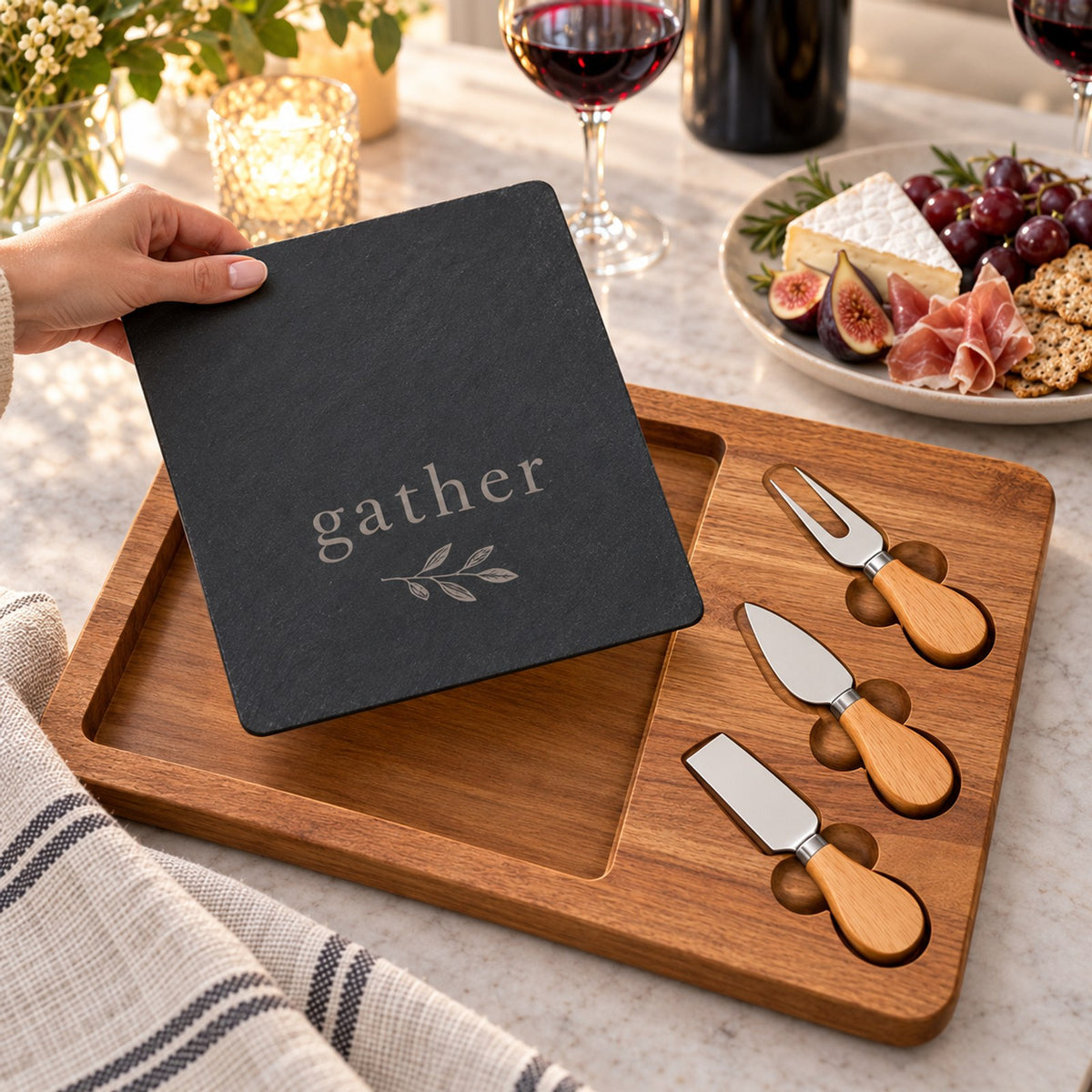 Black slate engraved with gather resting on an acacia board with three cheese knives, figs and wine on a candlelit table