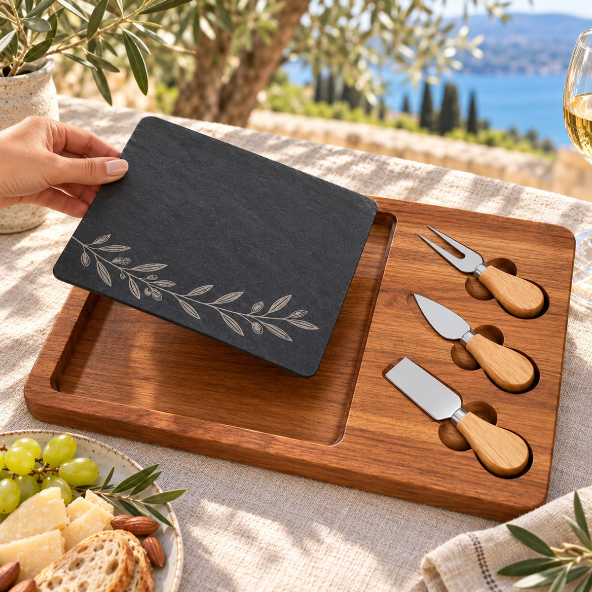 Black slate engraved with an olive branch on an acacia board with three cheese knives, on a sunny terrace table by the sea