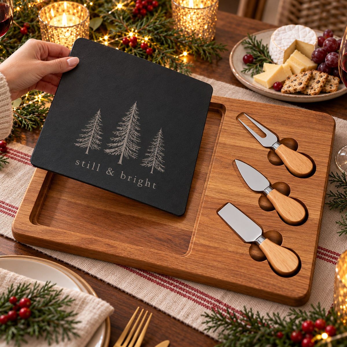 Black slate engraved with three evergreen trees and still & bright on an acacia board with cheese knives, on a Christmas table