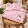 Pink bear-ear baby hooded towel with an embroidered gingerbread man, on a wooden stool with pine sprigs