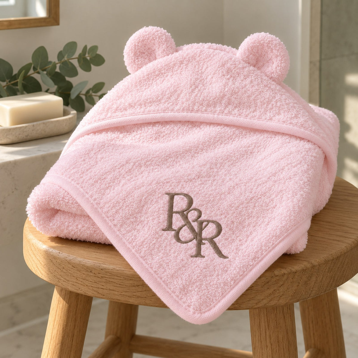 Pink bear-ear baby hooded towel with a taupe embroidered R&R monogram, folded on a wooden stool